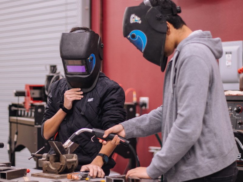 Two teenagers wearing welding face shields perform manufacturing work